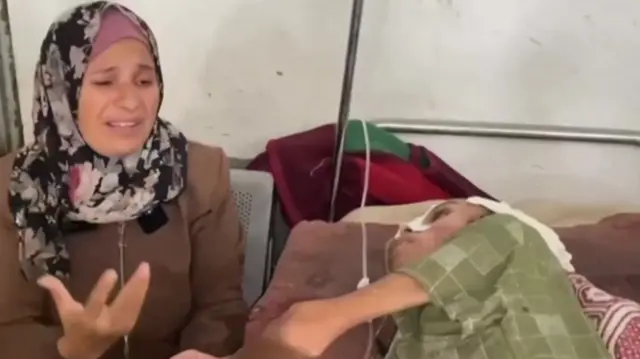 The mother, wearing a floral headscarf, gestures as she speaks, in tears. She holds the hand of her son.