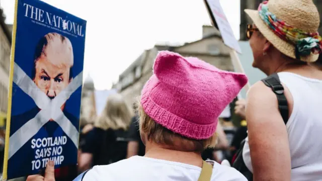 Two women pictured from the back attending an anti-Trump protest. The one at the centre, wearing a pink wool hat, holds a sign bearing a mock up front page of The National newspaper with the white saltire of the Scottish flag crossing over Donald Trump’s mouth