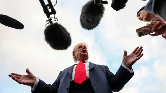 US President Donald Trump speaks to the media wearing a suit and red tie, gesturing with his hands.