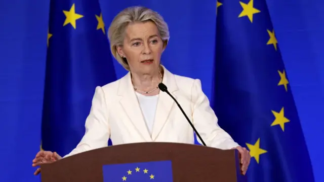 Ursula von der Leyen wearing a white blazer and white shirt stands at a wooden lectern bearing the EU flag. Two EU flags (bright blue with 12 yellow stars) stand behind von der Leyen