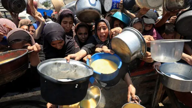 Palestinian women wait for food at a charity kitchen. They are holding out bowls towards the camera, some filled with what appears to be porridge. some are screaming.
