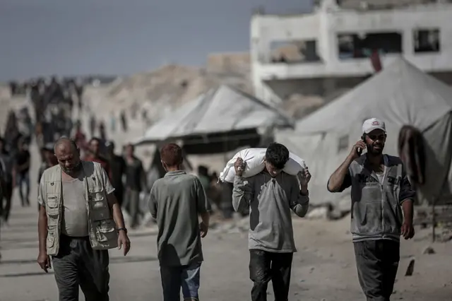 Palestinians collecting aid at distribution points in Gaza. The shells of ruined buildings are visible in the background and one young man is carrying a sack of aid.