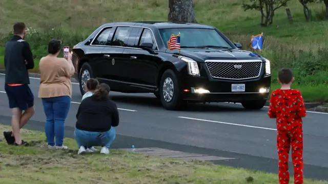 People line the sides of the motorway to watch President Trump pass. A woman crouches and a boy in red pyjamas watches the car.