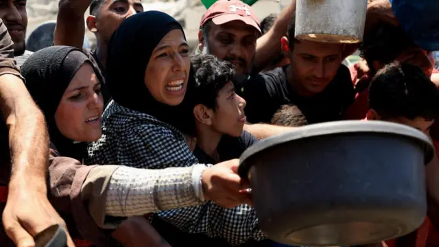 A woman, part of a big crowd, hold out a large bowl waiting for food at an aid point in Gaza.
