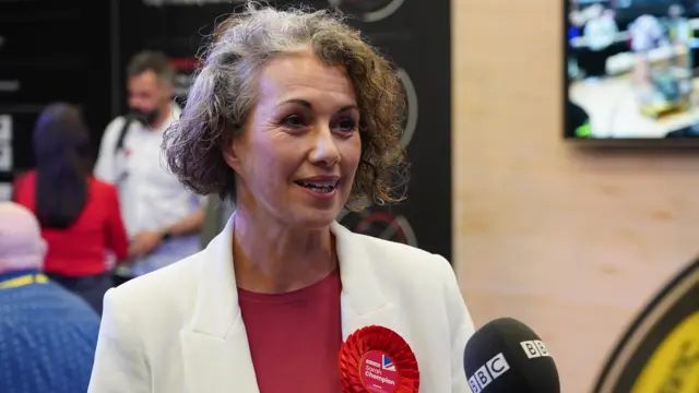 Sarah Champion gives interview to BBC in white blazer and red dress, people walking in the blurred background