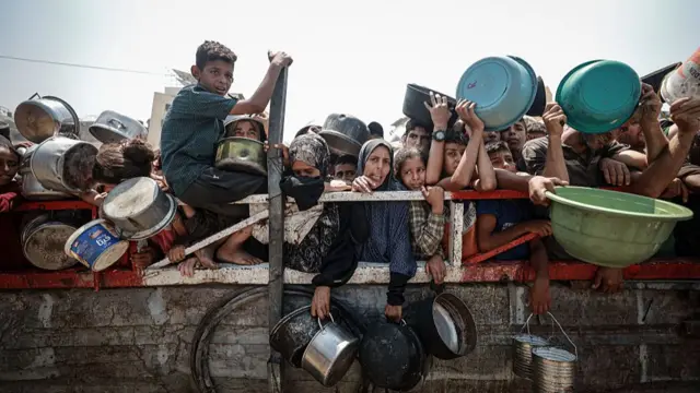Palestinians queuing outside a red and white metal barrier holding plastic basins and metal pots while waiting for food at a charity kitchen in Gaza City