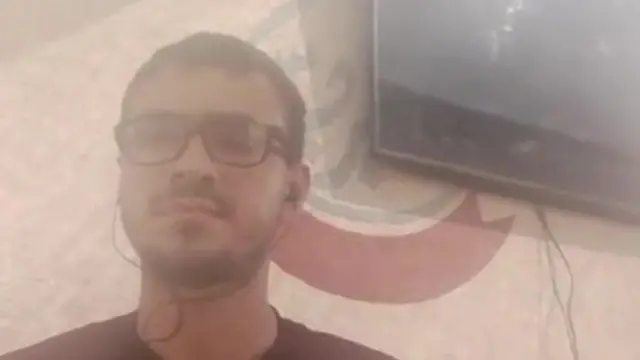 Screenshot of video call with a young man with glasses and black headphones. Image is grainy but there's a television hanging on the wall visible behind him