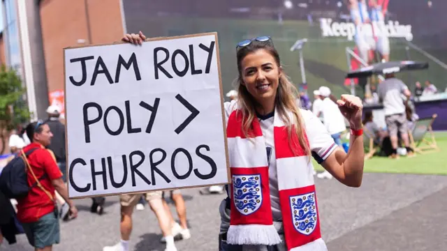 A fan of England holds up a sign which reads "Jam Roly poly > Churros" as they enjoy the pre-match atmosphere at the Fan Zone