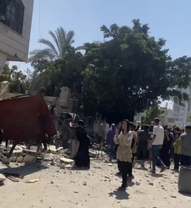 A screenshot shows palestinians walking in the area around rubble from an air strike in Gaza City