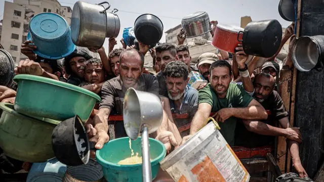 Large group of men pile up against a metal barrier as they reach for a ladle distributing food with plastic and metal pots and pans