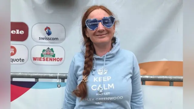 A woman wearing blue glasses and a blue sweatshirt smiles at the camera