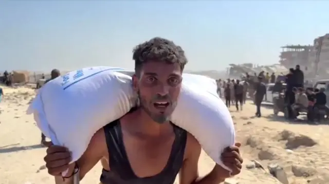 Mohammed al-Masry carries a white bag of aid on his shoulder among people and rubble