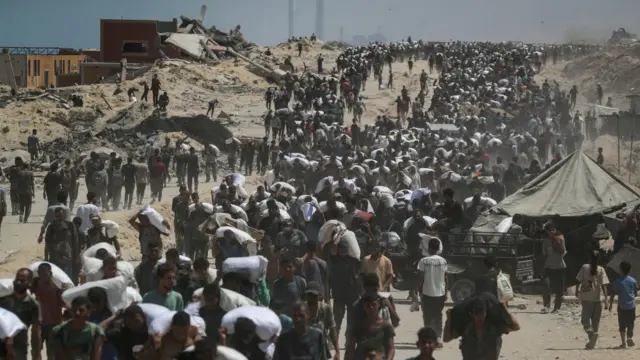 A large crowd makes it way along a route, with many carrying large white bags, which appears to have aid inside Israel backs down and pauses onslaught in parts of Gaza.