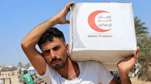 A man, who is looking at the camera, and is carrying a box of aid on his left shoulder that says "Emirates Red Crescent".