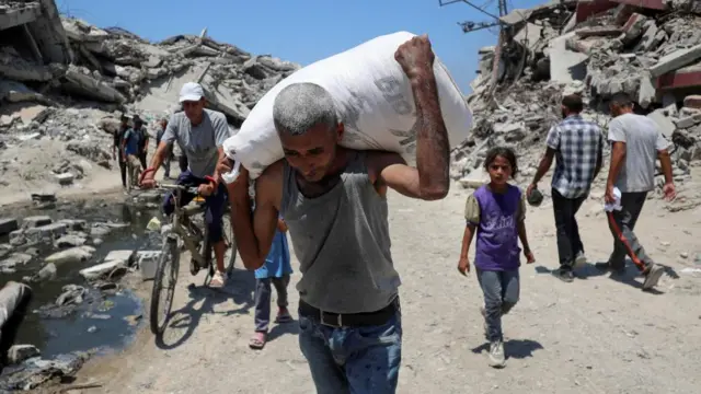 A man carries a white sack containing aid atop his shoulders as he walks don a street past ruins in Gaza