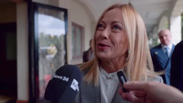 Meloni in grey blazer and white striped shirt smiling as she talks into two microphones