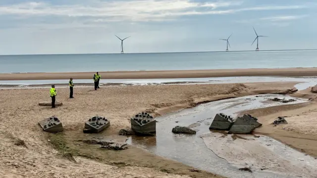 A beach looking out to sea, with police officers stood on the sand in a line. There's three wind turbines in the background far out in the sea