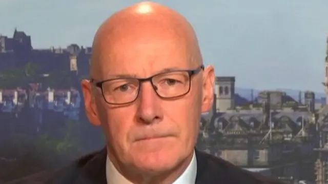 Headshot of Swinney who looks serious with his mouth pressed together. There's a backdrop of the view of Edinburgh and the castle that's obscured by Swinney's head