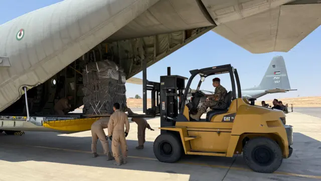 A forklift loads large parcels of aid onto a military plane