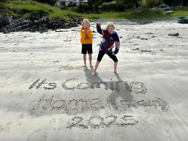 An image of two children with the words 'It's coming home (again) 2025' wrote in the sand.