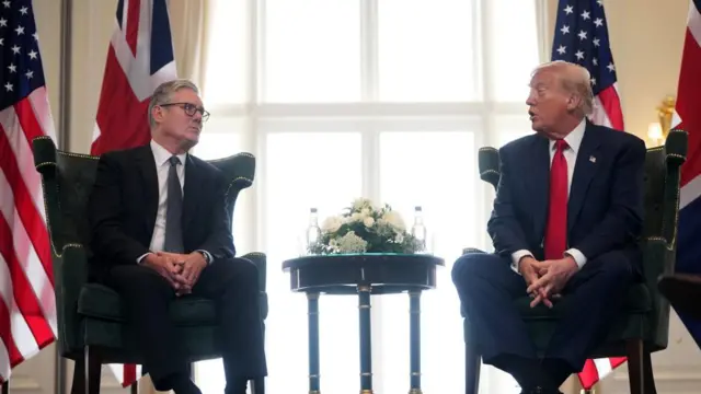 Starmer and Trump sitting on black suede chairs with gold details on them. There's a table in between with a floral arrangement and bottles of water. Behind them is a large window that is bright, and US and British flags too