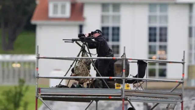 A sniper in all black looking through a view finder on top of a platform