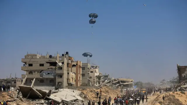 Pallets of aid are parachuted into Gaza, while a crowd of people on the ground walk along a route carrying bags of aid amid badly damaged buildings