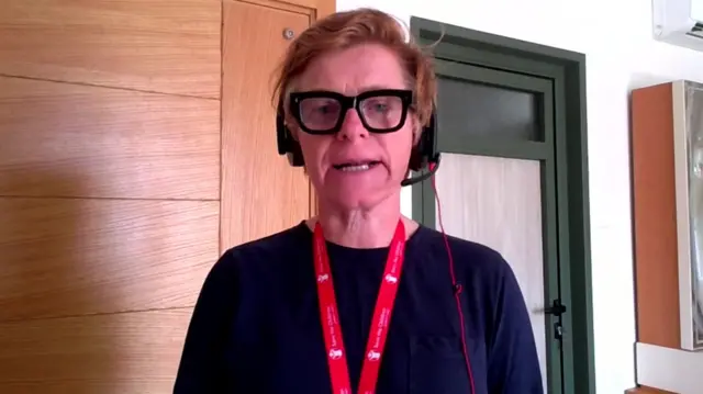Rachael Cummings sits in a room, wearing headphones and thick rimmed glasses, speaking to the camera
