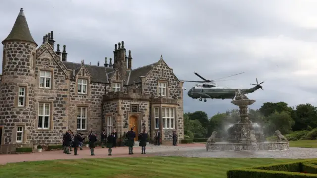 A helicopter is seen landing in front of a 19th century building, made of stone. There is a group of people out front, dressed in kilts and playing bagpipes.