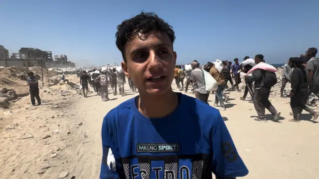 A man in a blue t shirt stands on a dusty road in Gaza, A large group of peoe