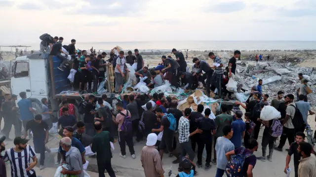 An empty truck is in the foreground with people climbing on top of it and standing around. Some carry white aid parcels