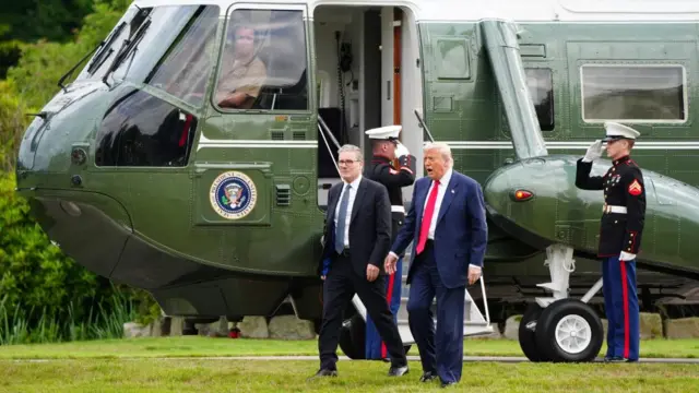 Trump and Starmer walking on the grass in front of a large green helicopter. Inside the pilot looks out the window at them. Two soldiers stand saluting him