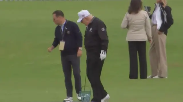 Trump on a golfing green holding a golf club in front of a basket of balls. People stand around him while he looks down