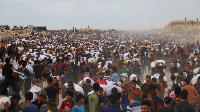 Scores of people are amassed in the picture, some carrying white parcels on their backs containing supplies