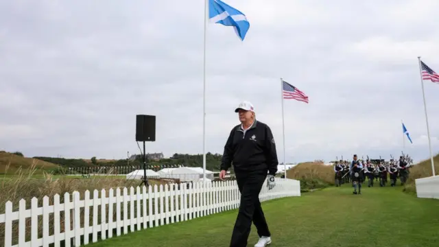 Trump walks along a grassy walkway lined by American flags and Saltires, and white fences. A Scottish band with bagpipers follows him