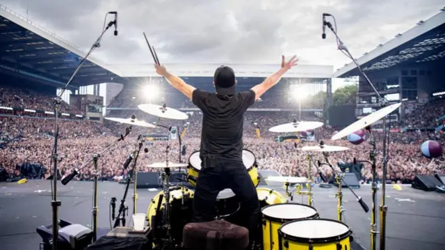 A man stands behind a drumkit on a stage and holds his arms up to a large crowd. He is wearing all black and a black baseball cap that is on backwards.