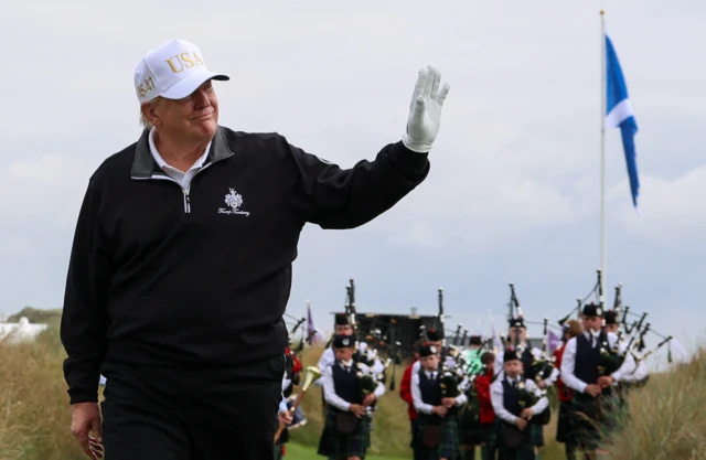 President Trump waving. He wears a black quarter zip sweater, a white polo shirt, a white glove and a white cap with USA written in gold. There is a group of bagpipe players behind him.