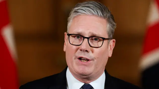 Keir Starmer addresses reporters at Downing Street. The photo is a close-up of his face - he's wearing glasses, a black suit and a white shirt.