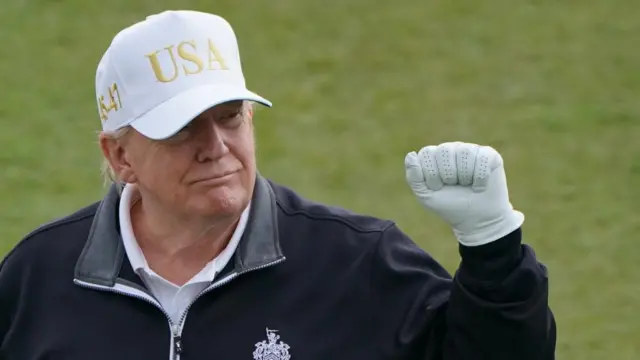 US President Donald Trump wearing a white cap with letters USA in gold writing. He is holding up his fist while wearing a white glove