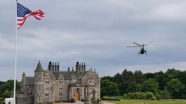 A helicopter landing o grass in front of a fancy old stone mansion with an American flag flying on a tall flag pole
