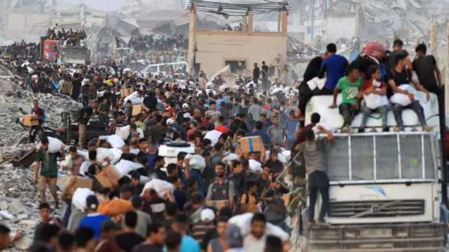 The image shows trucks trying to make their way down a road filled with people - some carrying white aid parcels. Some people sit on top of the trucks. Damaged infrastructure can also be seen