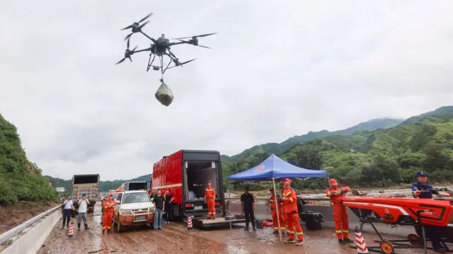 A drone carrying a bag hovering in the air, surrounded by rescuers in orange jumpsuits