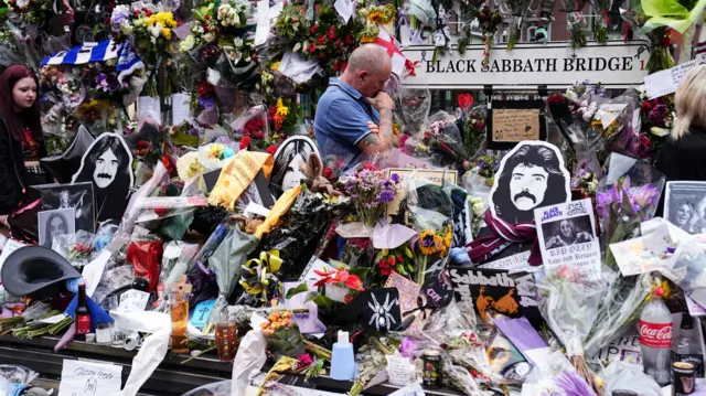 A man stood surrounded by flowers and notes and a sign saying Black Sabbath Bench