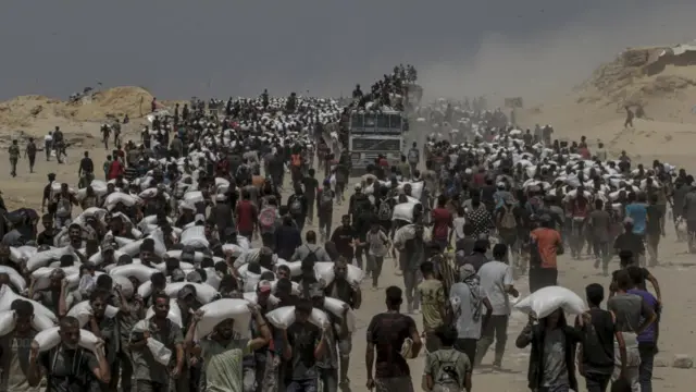 A crowd of people, mostly men, surround a lorry, many walking away with large white sacks across their shoulders