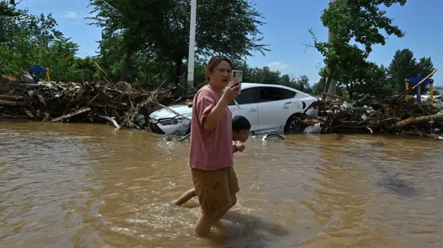 A woman knee-deep in water, walking with her child. She is holding up her phone.