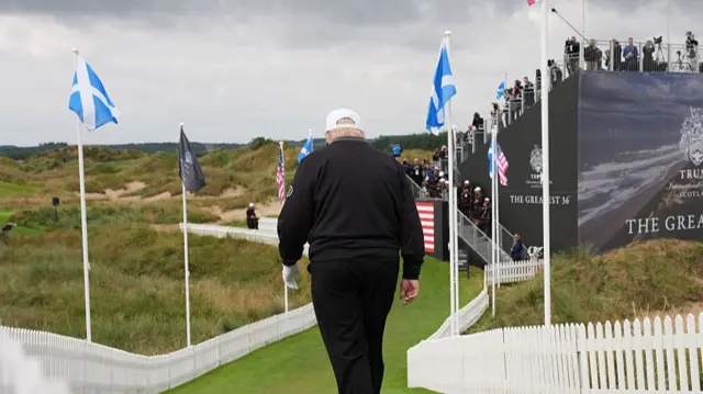 Trump walking away from the camera along a grassy path with Scottish and American flags
