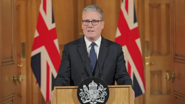 Keir Starmer stoof behind a podium, giving a statement. There are two Union Jack flags behind him.
