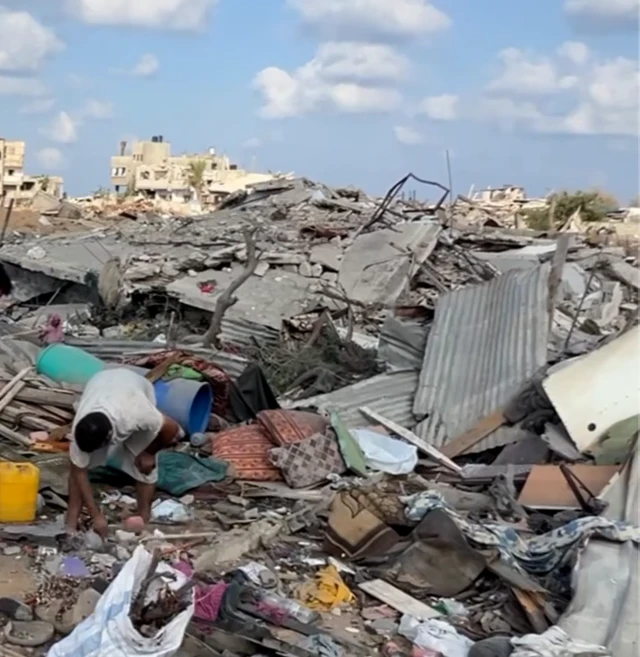 A still image from a video. It shows a destroyed building, with a man bending down while standing on the rubble.