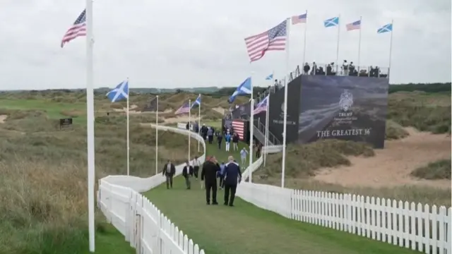 People walking along a path in between two fence lines in front of a golf course. There are American and Scottish flags lining the path.