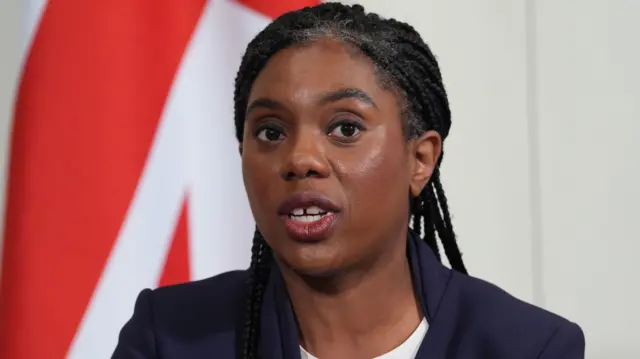 Kemi Badenoch speaks to the media; she's wearing a blue suit and white top, and she's standing in front of a union flag.